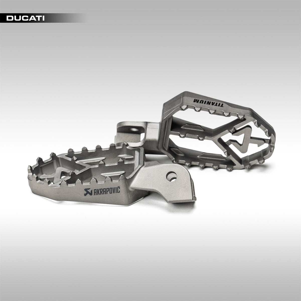 Titanium foot pegs with 'Akrapovic' branding on a gray background
