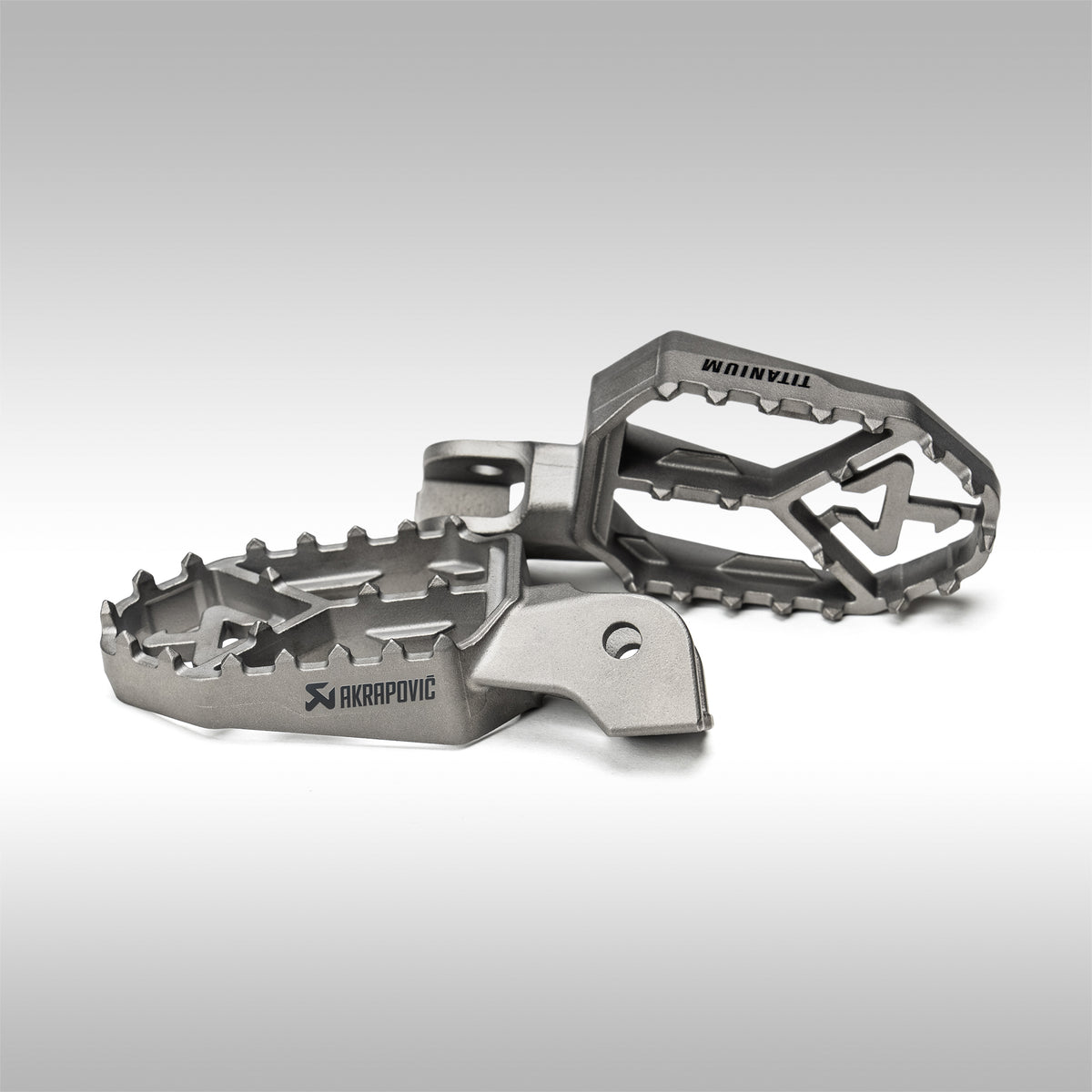 Titanium foot pegs with 'AKRAPOVIC' branding on a gray background