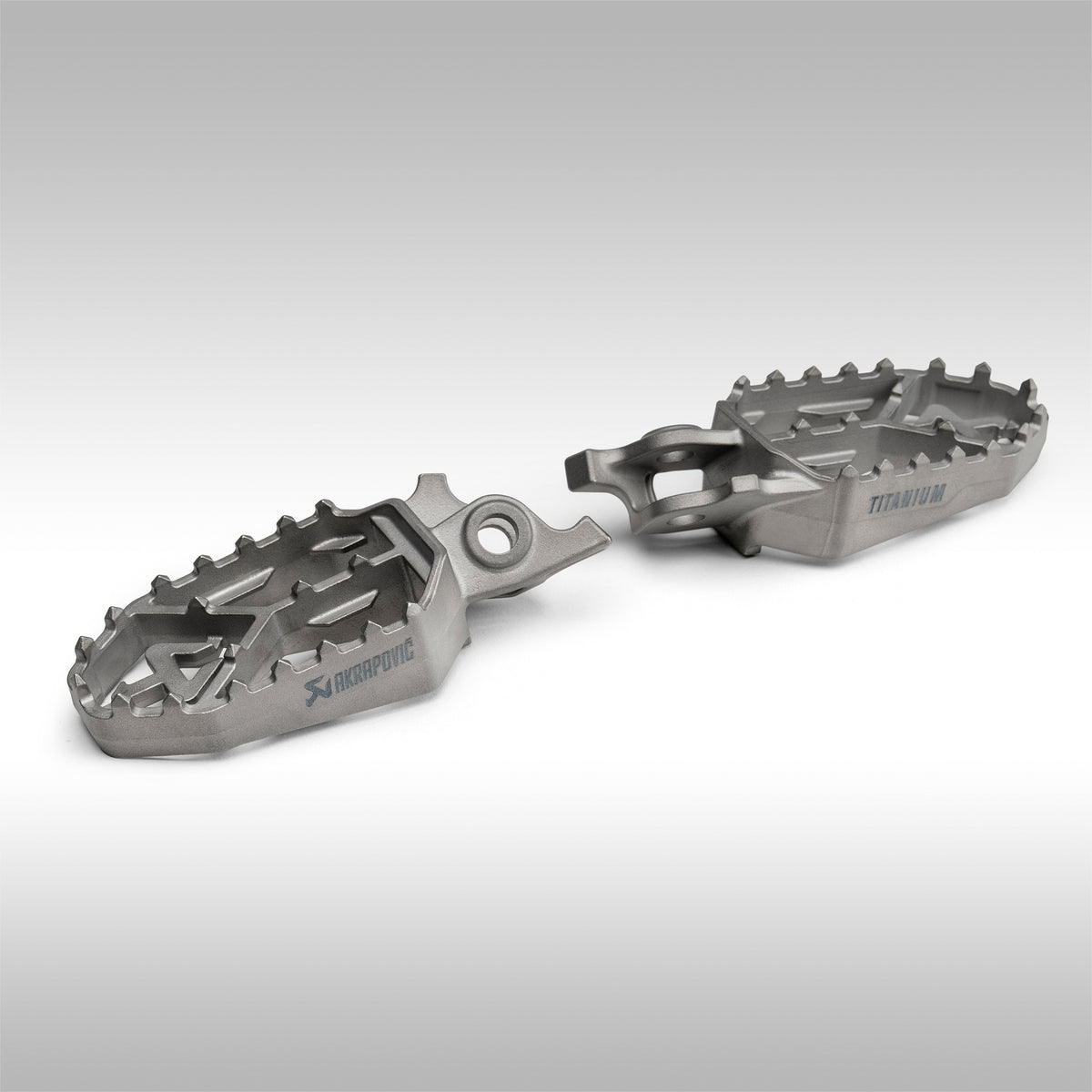 Two Akrapovic titanium foot pegs on a gray background.