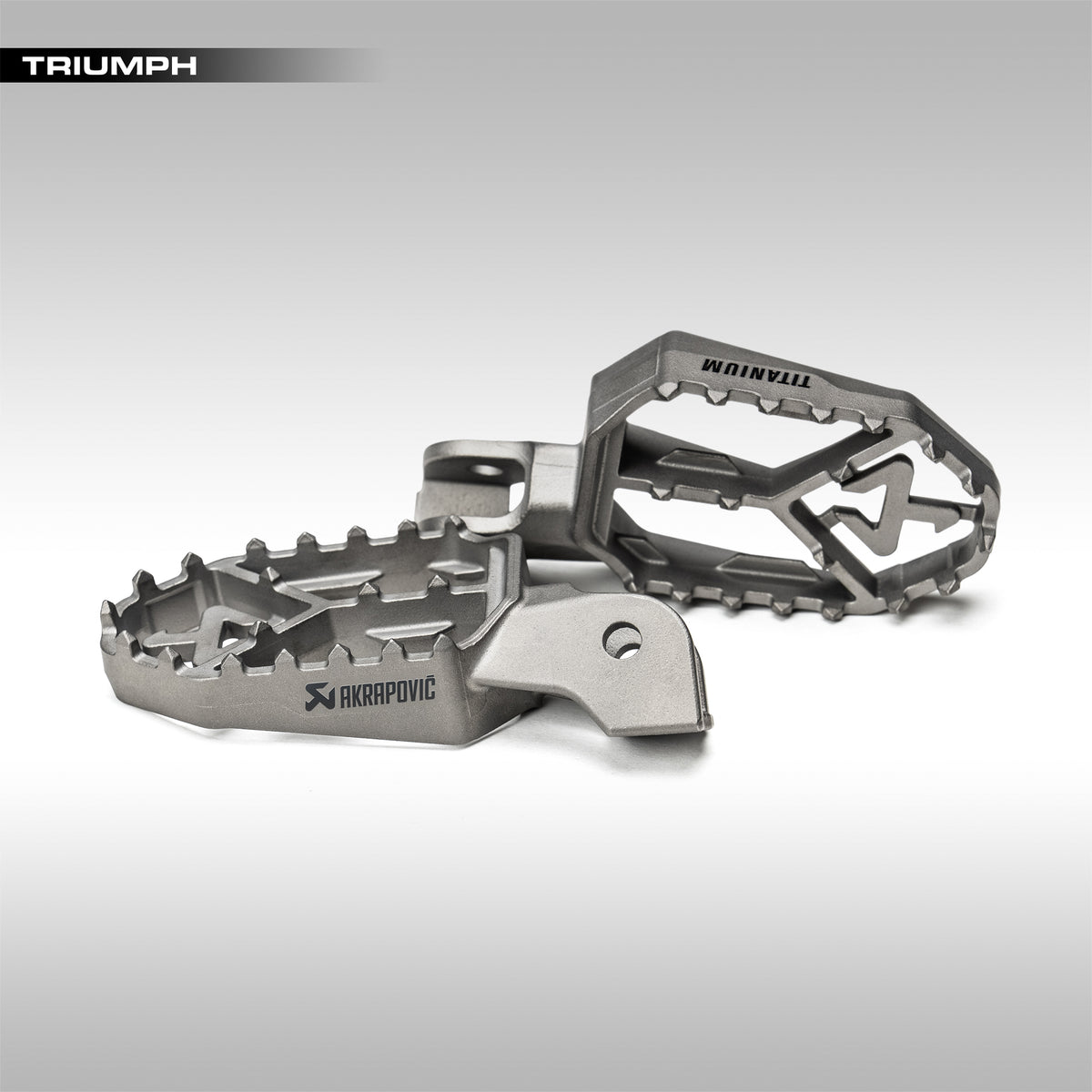 Two Akrapovic titanium foot pegs for Triumph Tiger on a gray background.