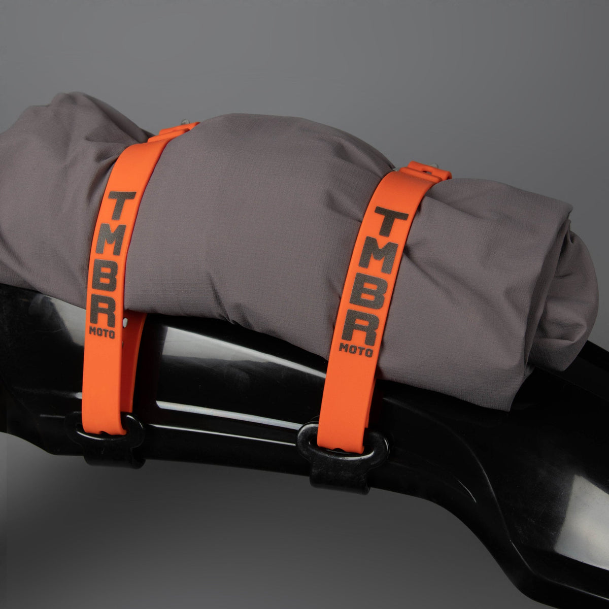 Orange TMBR MOTO fender strap kit holding a gray jacket to a black fender